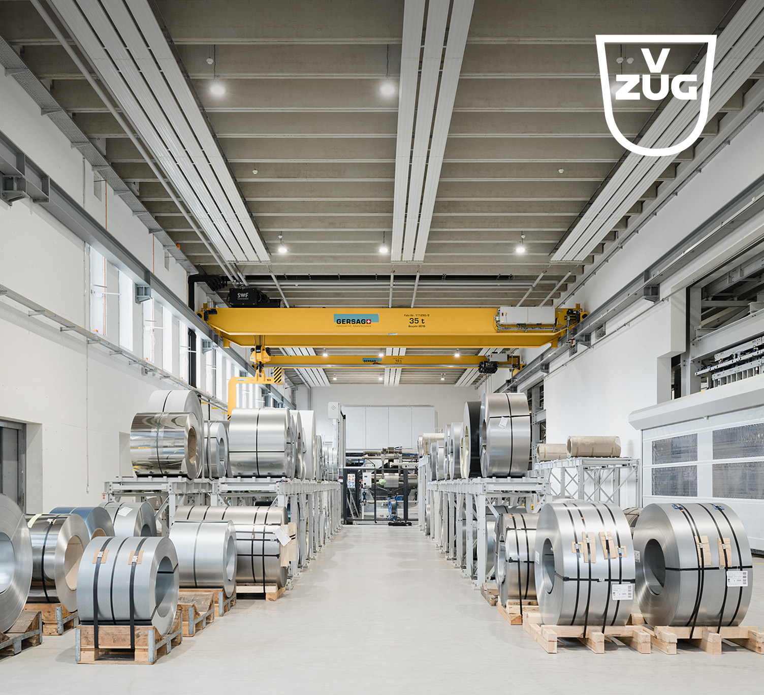 Produktionshalle der V-ZUG mit auf Paletten gelagerten Stahlcoils und einem gelben Deckenkran – moderner Industriearbeitsplatz für Haushaltsgerätefertigung.
