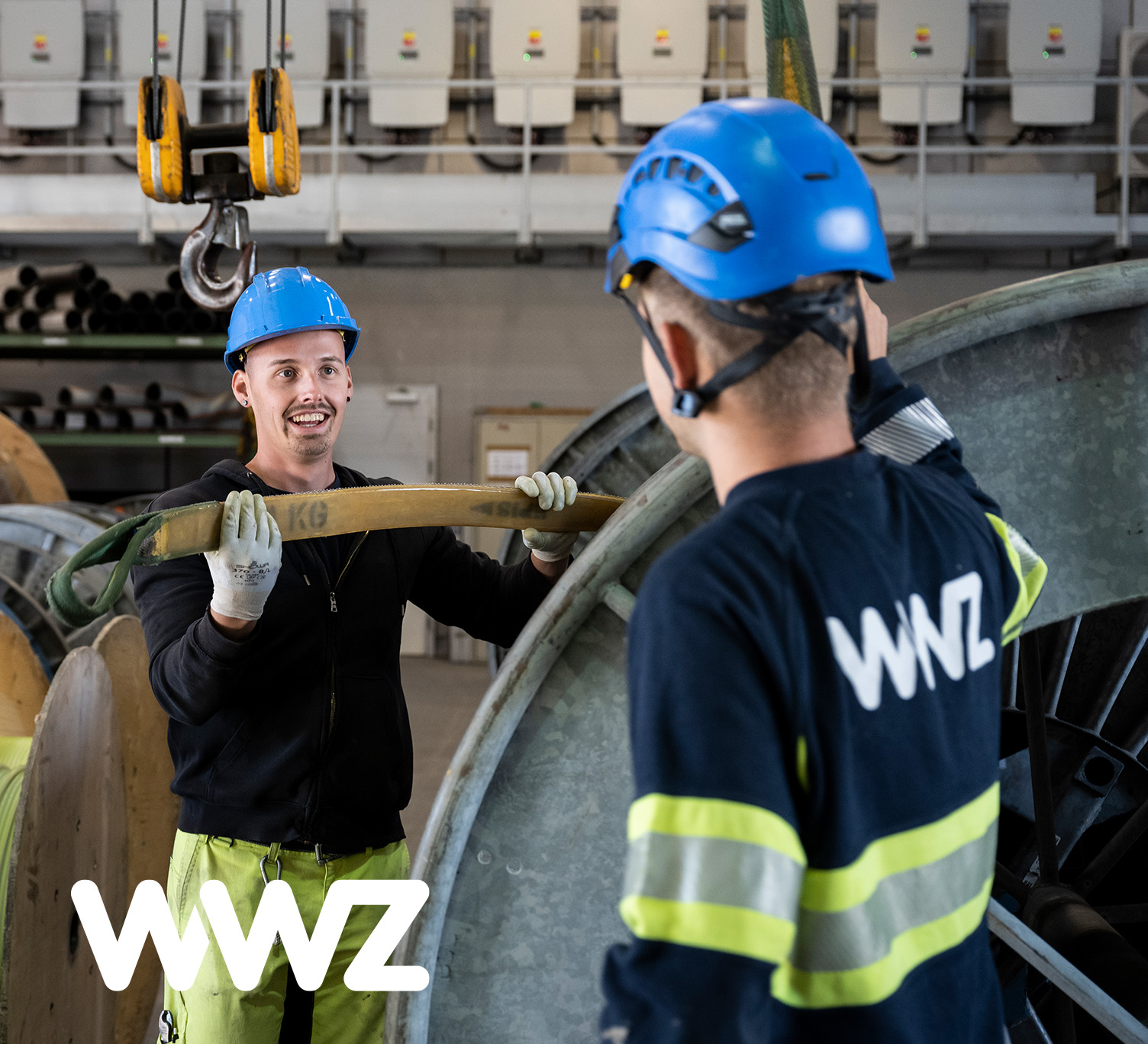 Zwei Mitarbeitende der WWZ mit blauen Helmen und Sicherheitskleidung bei der Arbeit an einer grossen Kabeltrommel in einer Industriehalle.