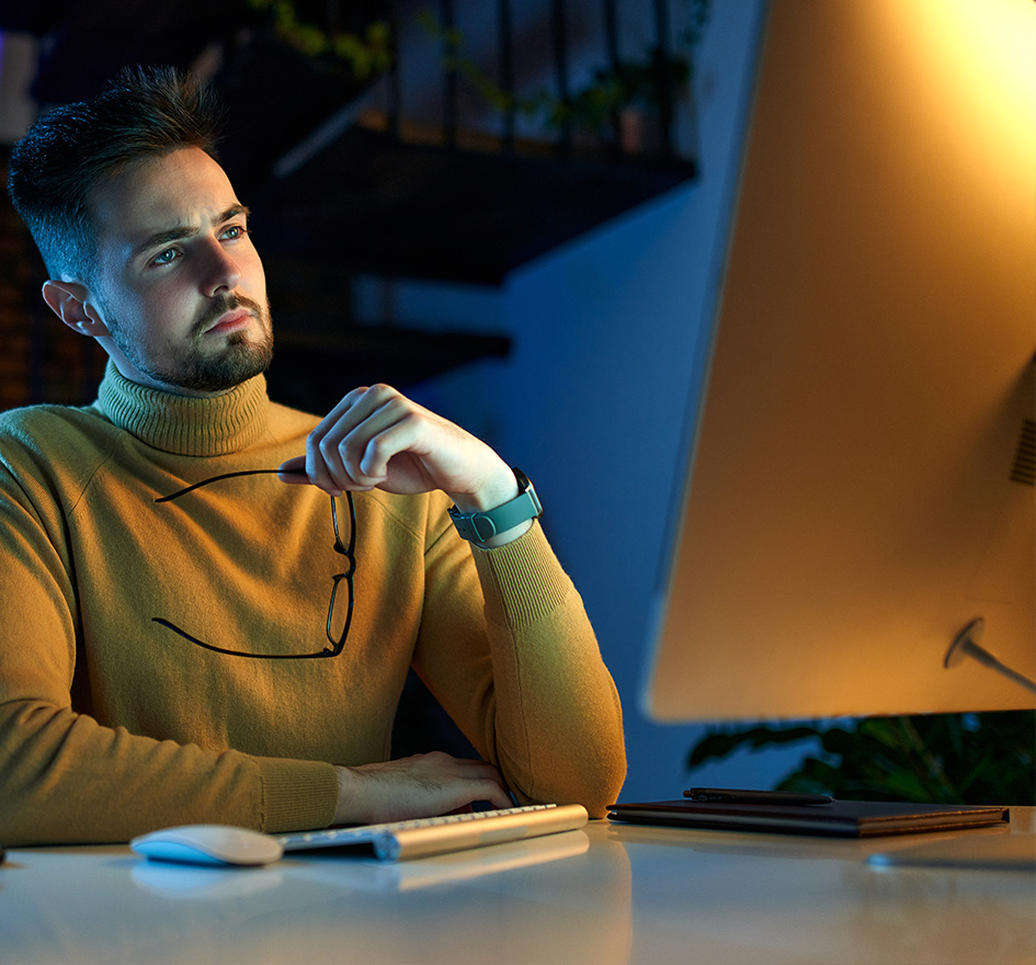 Nachdenklicher Mann in Pullover sitzt am Schreibtisch mit Computer und hält nachdenklich seine Brille – symbolisiert Abhängigkeit und Entscheidungsdruck in Bezug auf IT-Anbieter.