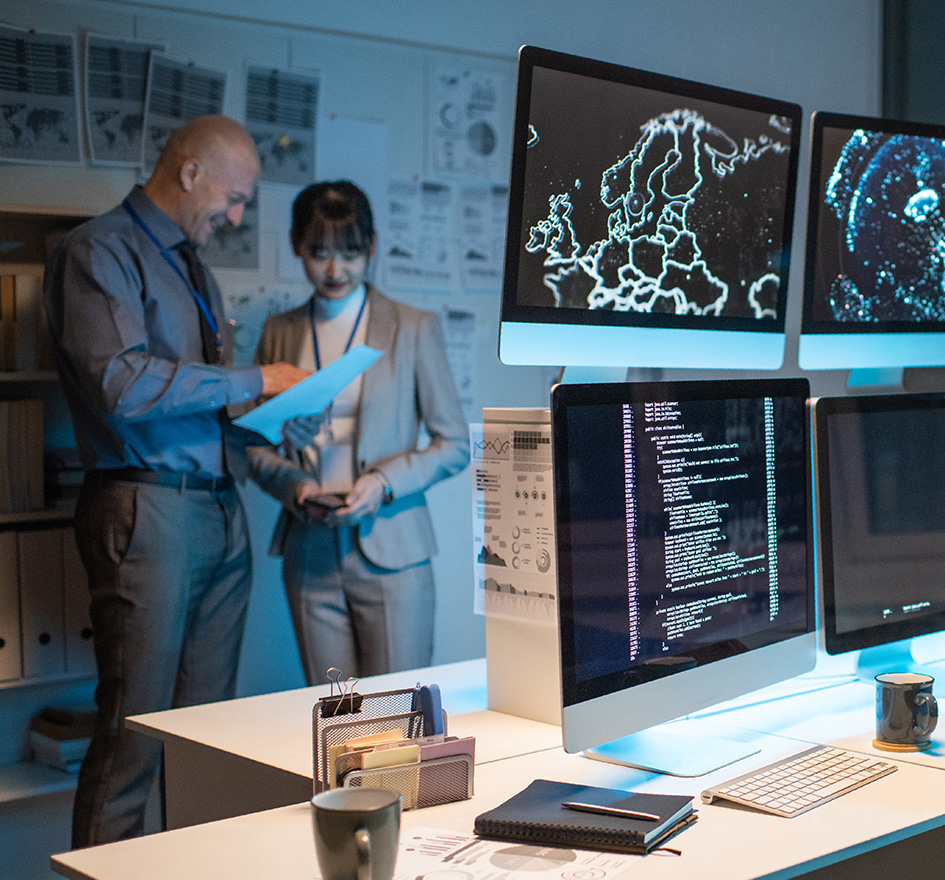 Zwei Personen in einem Technikbüro mit Monitoren, die Code und Karten anzeigen – symbolisiert hohe IT-Komplexität, Wartungsaufwand und technischen Betreuungsbedarf.