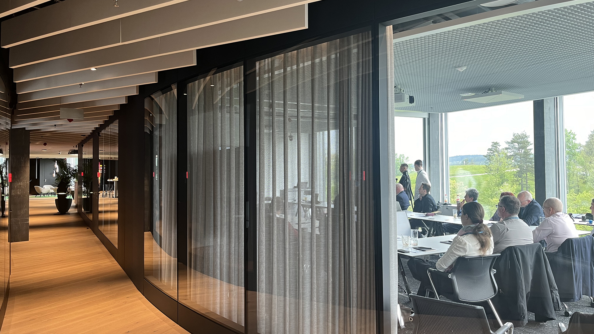 Modernes Bürogebäude mit gläsernen Besprechungsräumen und Sicht auf einen laufenden Event mit Teilnehmenden, im Hintergrund grüne Landschaft.