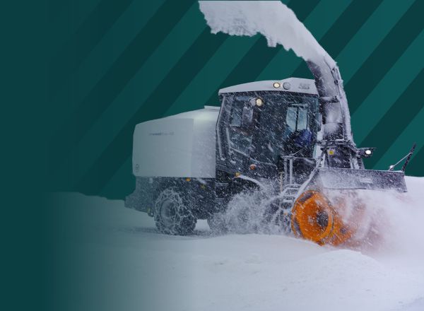 Schneefräse mit orangem Fräskopf räumt bei Schneefall eine verschneite Strasse und wirft den Schnee in hohem Bogen aus
